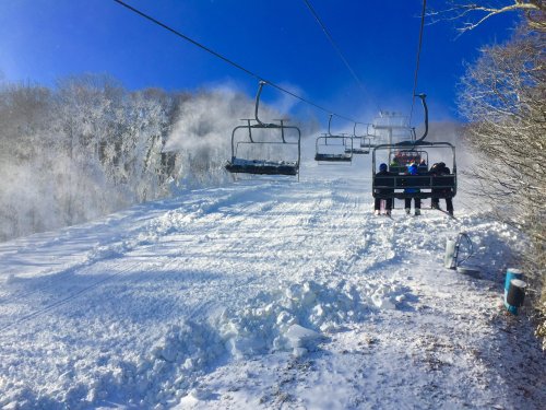 Ski lift chairs ascend a snowy slope under a clear blue sky, with snow blowing in the air.