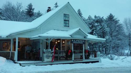 A quaint grocery store with a snowy exterior, decorated with wreaths, surrounded by trees in a winter landscape.