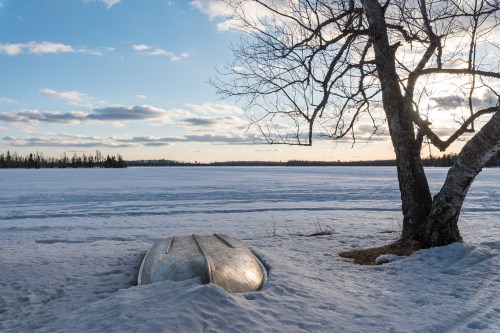 A snowy landscape with a boat resting on the ground beside a bare tree, under a blue sky with scattered clouds.