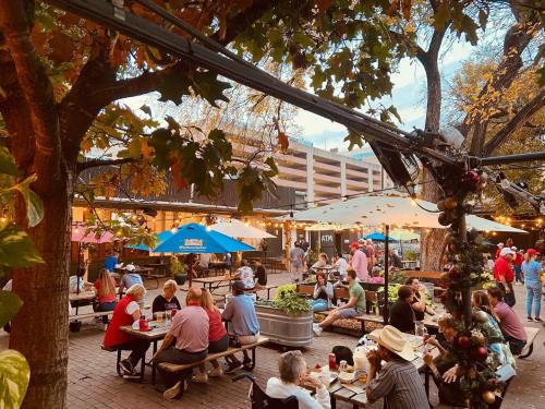 A lively outdoor dining area with people at tables, string lights, and colorful umbrellas among trees and greenery.