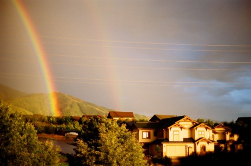 A vibrant double rainbow arcs over a hillside, with houses and trees in the foreground under a cloudy sky.