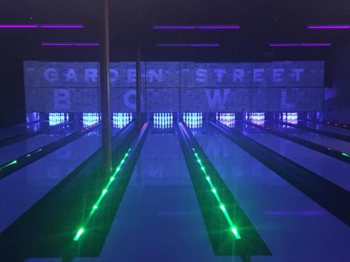 A bowling alley with illuminated lanes and a backdrop displaying "Garden Street Bowl" in neon lights.