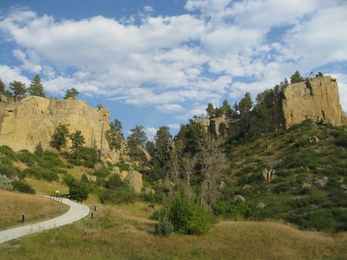 A winding path leads through a rocky landscape with cliffs and trees under a blue sky with scattered clouds.