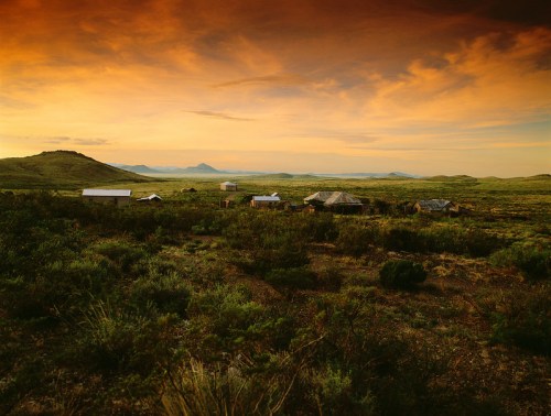 A serene landscape at sunset, featuring a small cluster of buildings surrounded by lush greenery and distant mountains.