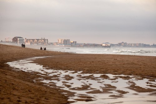 A sandy beach with gentle waves, people walking in the distance, and a pier and buildings along the shoreline.