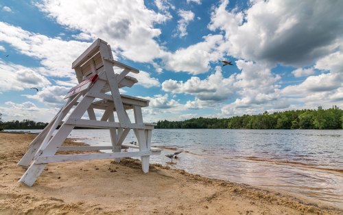 A lifeguard chair on a sandy beach by a calm lake, with clouds and birds in the sky.