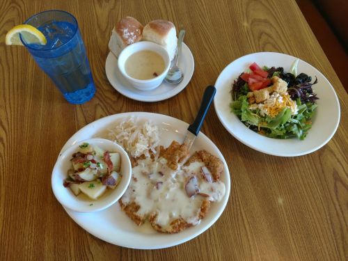 A plate with chicken fried steak topped with gravy, sides of coleslaw, salad, and a bowl of soup, with rolls and a drink.