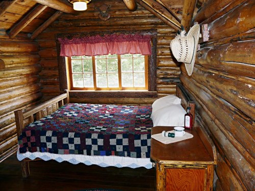 Cozy log cabin bedroom with a patchwork quilt, wooden furniture, and a window letting in natural light.