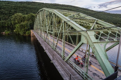 A green metal bridge spans a river, with two motorcycles riding across and lush hills in the background.