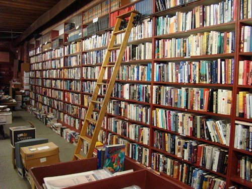 A wooden ladder leans against tall bookshelves filled with various books, with boxes on the floor below.