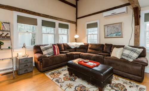 Cozy living room with a brown sectional sofa, decorative pillows, a coffee table, and large windows with natural light.