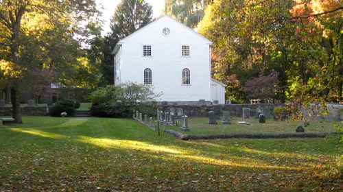 A white building surrounded by trees and gravestones in a serene, autumn landscape.