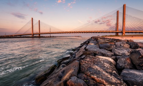 A modern suspension bridge spans a river at sunset, with rocky shoreline in the foreground and colorful sky above.