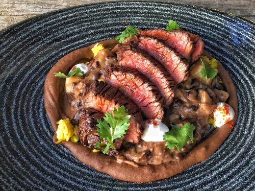 A plate of sliced steak atop a bed of beans, garnished with herbs and colorful toppings on a textured black dish.