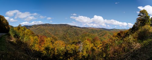 A panoramic view of colorful autumn foliage covering rolling mountains under a bright blue sky with fluffy clouds.
