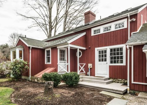 A charming red wooden house with a porch, surrounded by greenery and trees on a cloudy day.