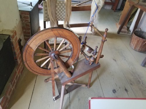 A wooden spinning wheel on a wooden floor, with a rustic background featuring woven materials and a basket.
