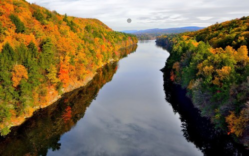 A serene river flows between vibrant autumn-colored hills under a cloudy sky. Reflections shimmer on the water's surface.