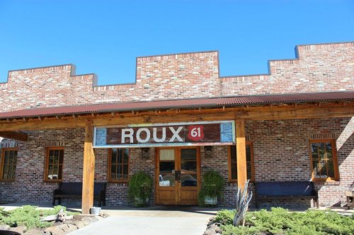 Exterior of Roux 61 restaurant, featuring a brick facade, wooden beams, and a sign above the entrance. Clear blue sky.