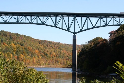 A large steel bridge spans a river, surrounded by colorful autumn foliage on the hillsides. Clear blue sky above.