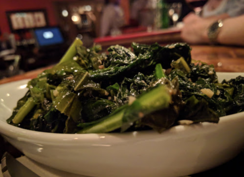 A close-up of a bowl of sautéed greens, featuring dark leafy vegetables garnished with sesame seeds.