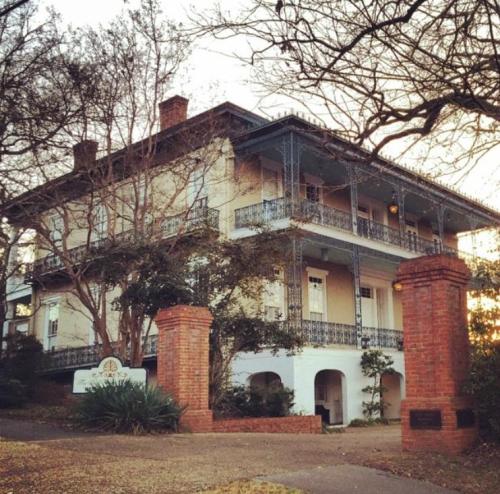 Historic three-story house with a wraparound porch, surrounded by trees and brick pillars at the entrance.