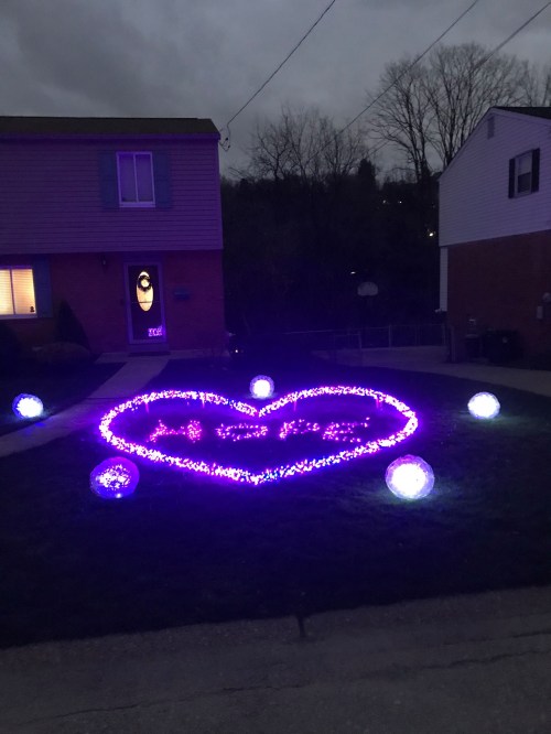 A yard decorated with purple lights arranged in a heart shape, surrounded by glowing orbs, in front of a house.