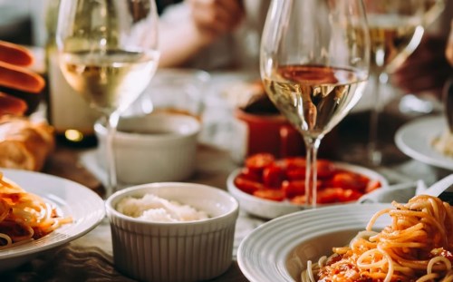 A table set with plates of pasta, bowls of rice and tomatoes, and glasses of white wine, creating a cozy dining atmosphere.