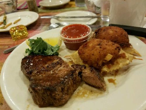 A plate featuring a grilled steak, crab cakes, a side of sauce, and garnishes of lemon and lettuce.