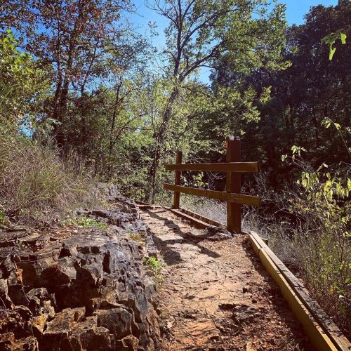 A winding path through a wooded area, bordered by rocks and a wooden fence, under a clear blue sky.