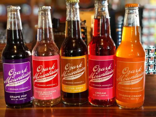 Five colorful bottles of soda from Ozark Mountain Bottleworks, featuring grape pop, lemonade, butter beer, cream, and orange cream.