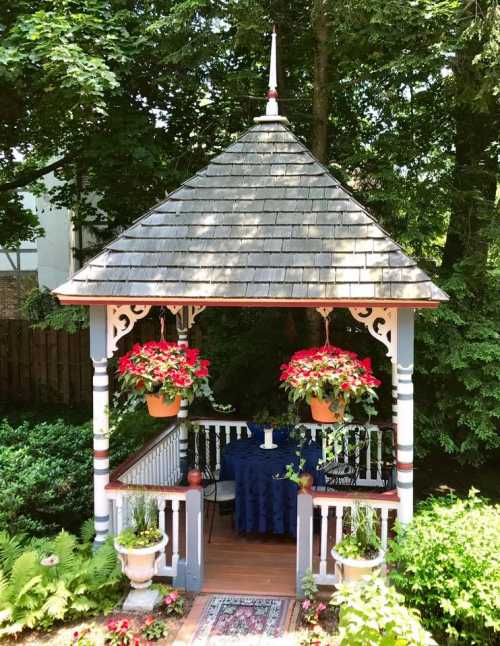 A charming gazebo surrounded by greenery, featuring hanging flower pots and a small table set with a blue tablecloth.