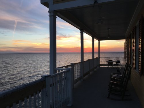 A serene sunset over the water, viewed from a porch with rocking chairs and white railings.