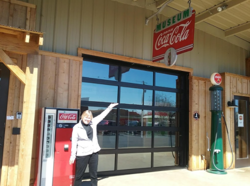 A woman stands outside a Coca-Cola museum, pointing at the entrance with vintage soda machines nearby.