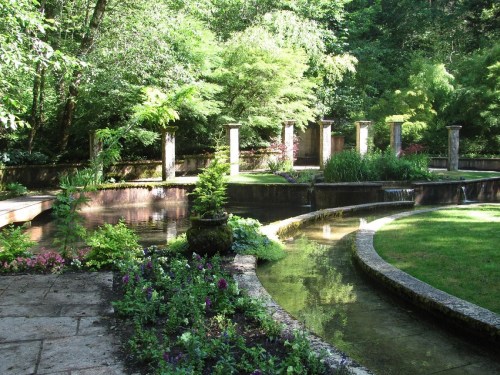 A serene garden with a pond, lush greenery, and stone pillars surrounded by colorful flowers and plants.
