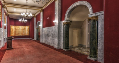 Elegant hallway with red walls, marble accents, and ornate chandeliers, featuring arched doorways and a plush carpet.