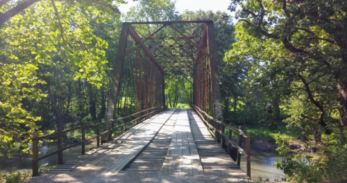 A rustic metal bridge spans a calm creek, surrounded by lush green trees and sunlight filtering through the leaves.