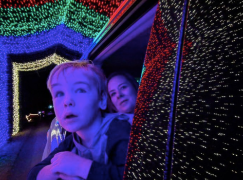 A child and an adult look out of a car window, surrounded by colorful holiday lights.