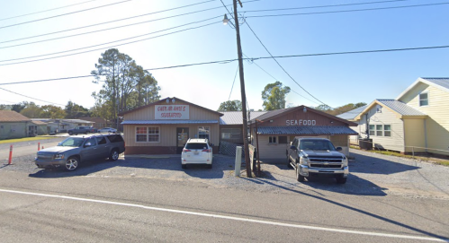 A seafood restaurant with a sign, featuring parked cars and a gravel lot, set along a roadside.