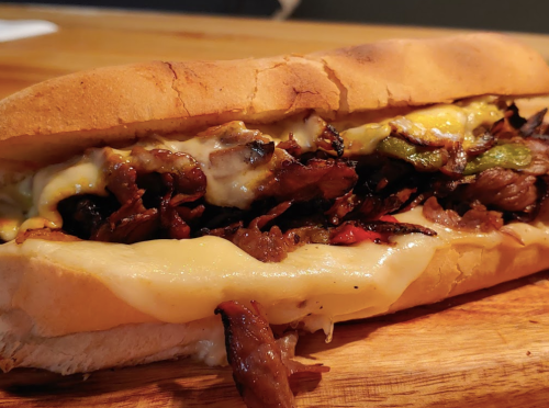 A close-up of a loaded sandwich with grilled meat, melted cheese, and peppers on a toasted hoagie roll.