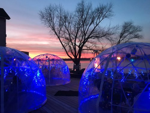 Two illuminated igloos on a deck by a lake at sunset, with vibrant colors in the sky and bare trees in the background.