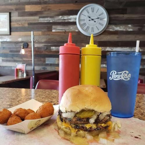 A juicy burger with pickles and onions, served with tater tots, ketchup, mustard, and a blue drink in a diner setting.