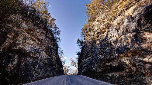 A narrow road between towering rocky cliffs, with trees and a clear blue sky above.