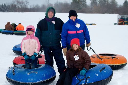 A family poses in winter gear with snow tubes on a snowy hill, smiling in a fun outdoor setting.