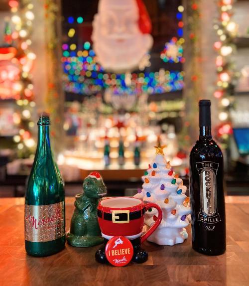 A festive display featuring a green bottle, a dinosaur figurine, a Christmas tree, a Santa mug, and a wine bottle.