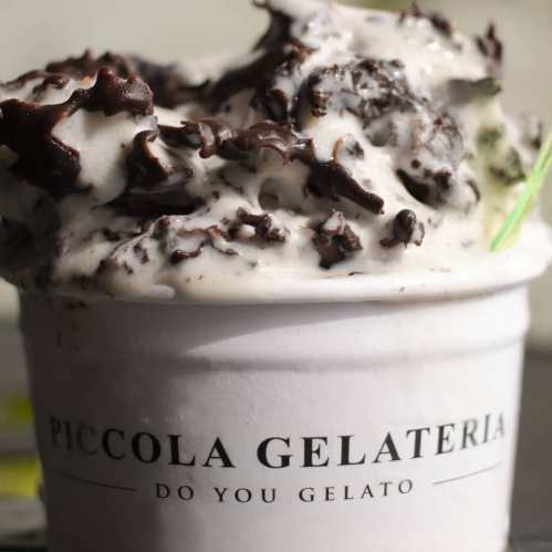 A close-up of a cup of gelato topped with chocolate flakes, featuring the label "Piccola Gelateria."