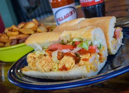 A sandwich filled with fried chicken, lettuce, and tomato, served with a side of potato wedges and drinks in the background.