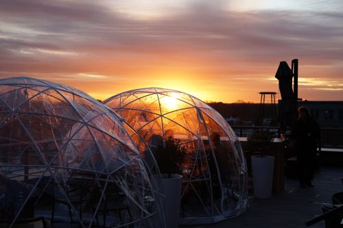 Two transparent igloos on a rooftop at sunset, with a vibrant sky and a silhouette of a person in the background.