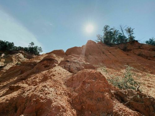 A sunlit rocky hillside with trees at the top, showcasing layers of orange and brown earth against a clear blue sky.