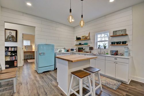 Bright kitchen with white shiplap walls, a blue retro fridge, wooden island, and open shelving displaying dishware.